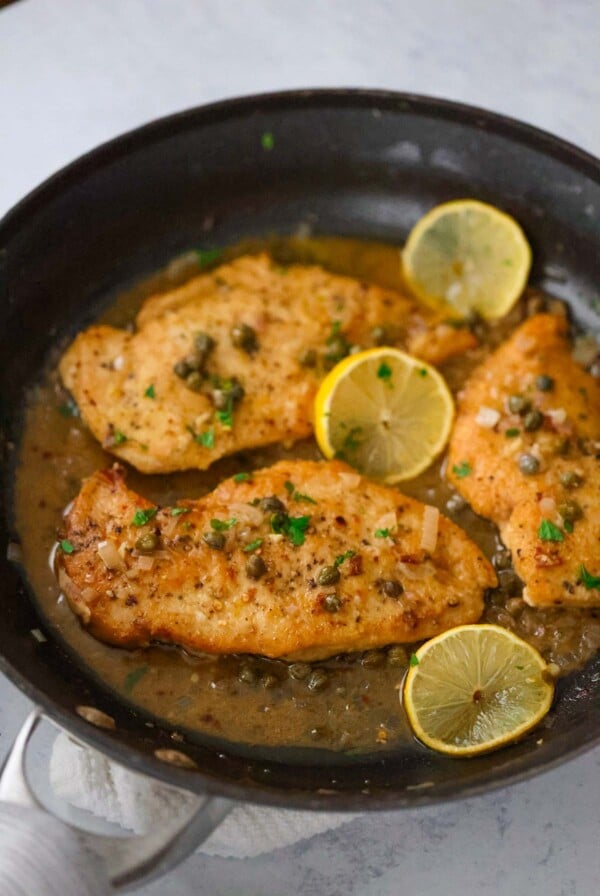 three chicken breasts piccata in a skillet with sauce and lemon slices.