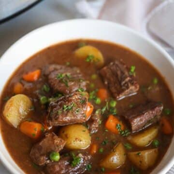 Beef stew in a white bowl with a crockpot in the background.