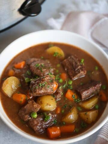 Beef stew in a white bowl with a crockpot in the background.