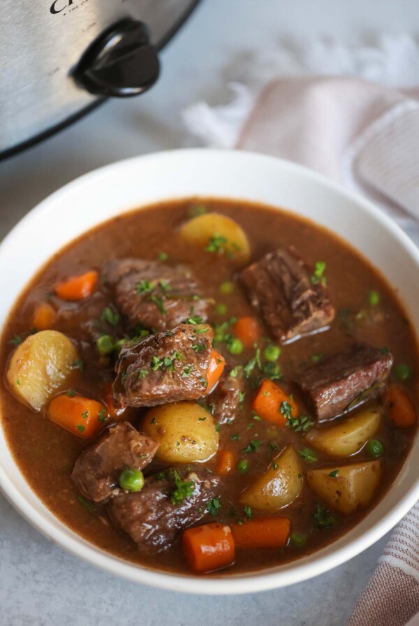 Beef stew in a white bowl with a crockpot in the background.