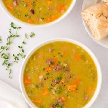 Two small bowls filled with split pea soup with ham and herbs and bread on the side.