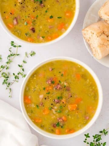 Two small bowls filled with split pea soup with ham and herbs and bread on the side.