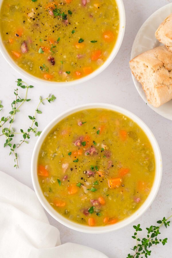 Two small bowls filled with split pea soup with ham and herbs and bread on the side.