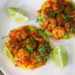 Two shrimp tostadas on a white plate with lime wedges on the side.