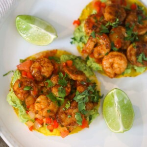 Two shrimp tostadas on a white plate with lime wedges on the side.