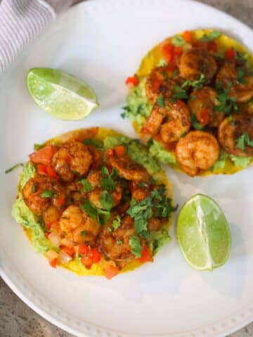 Two shrimp tostadas on a white plate with lime wedges on the side.