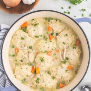 A pot filled with chicken and dumplings with onions, parsley, and garlic on the side.