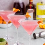 Three cosmopolitan martinis with alcohol bottles in the background.