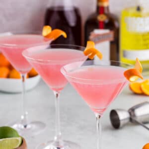 Three cosmopolitan martinis with alcohol bottles in the background.
