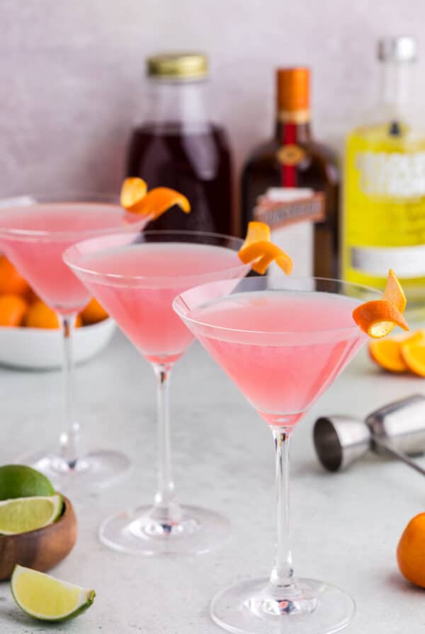 Three cosmopolitan martinis with alcohol bottles in the background.