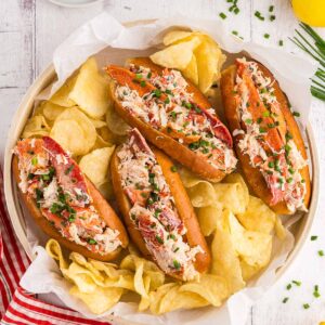 Four lobster rolls with chips, pickles, and lemon wedges.