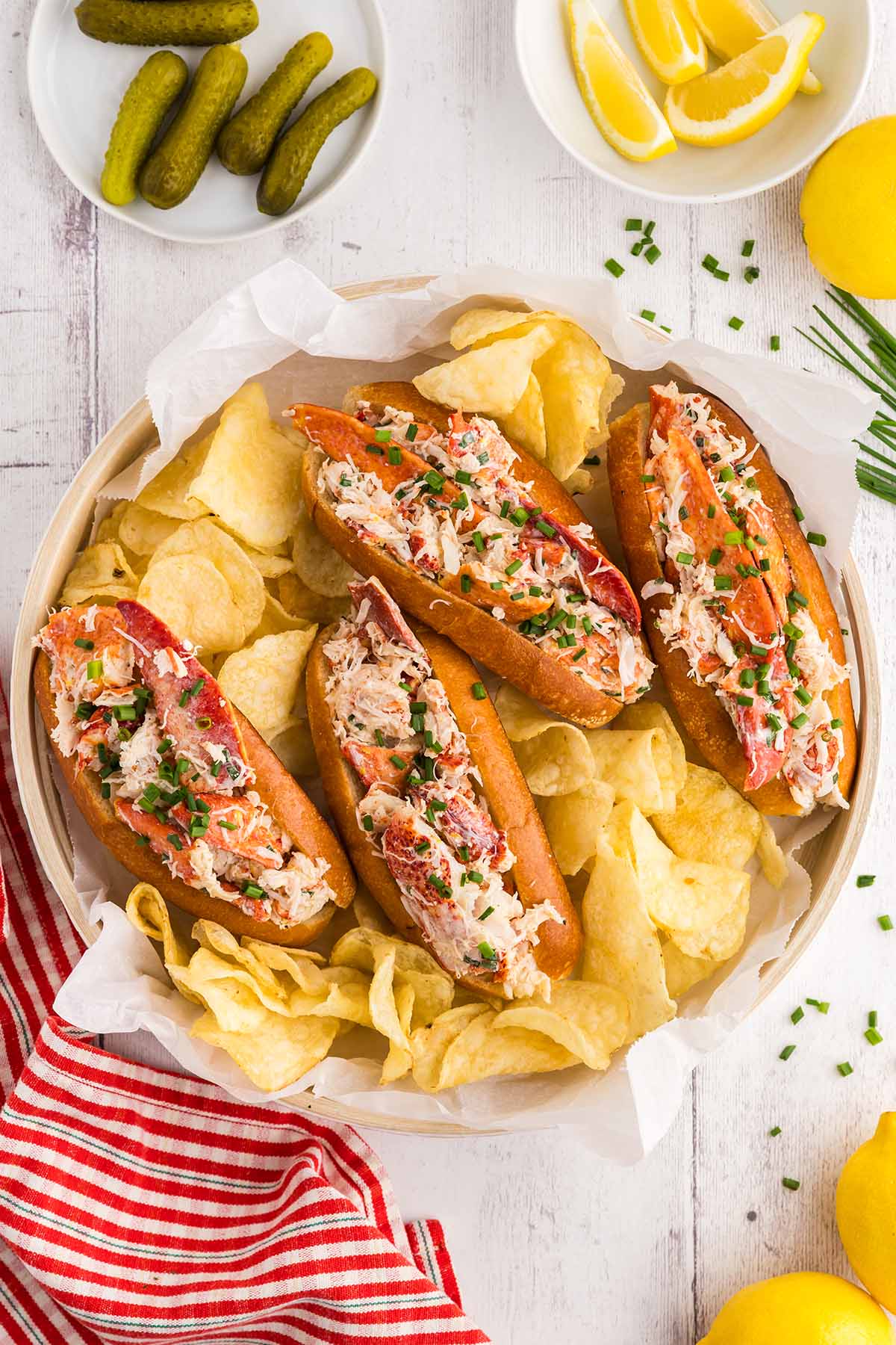 Four lobster rolls with chips, pickles, and lemon wedges.