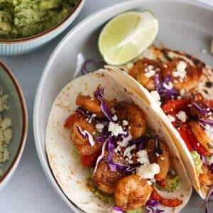 Two shrimp tacos with guacamole, limes, and cotija cheese on the side.