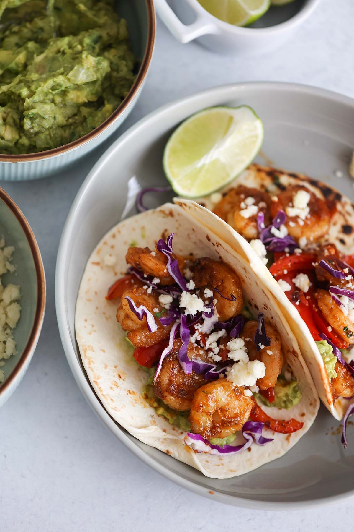 Two shrimp tacos with guacamole, limes, and cotija cheese on the side.