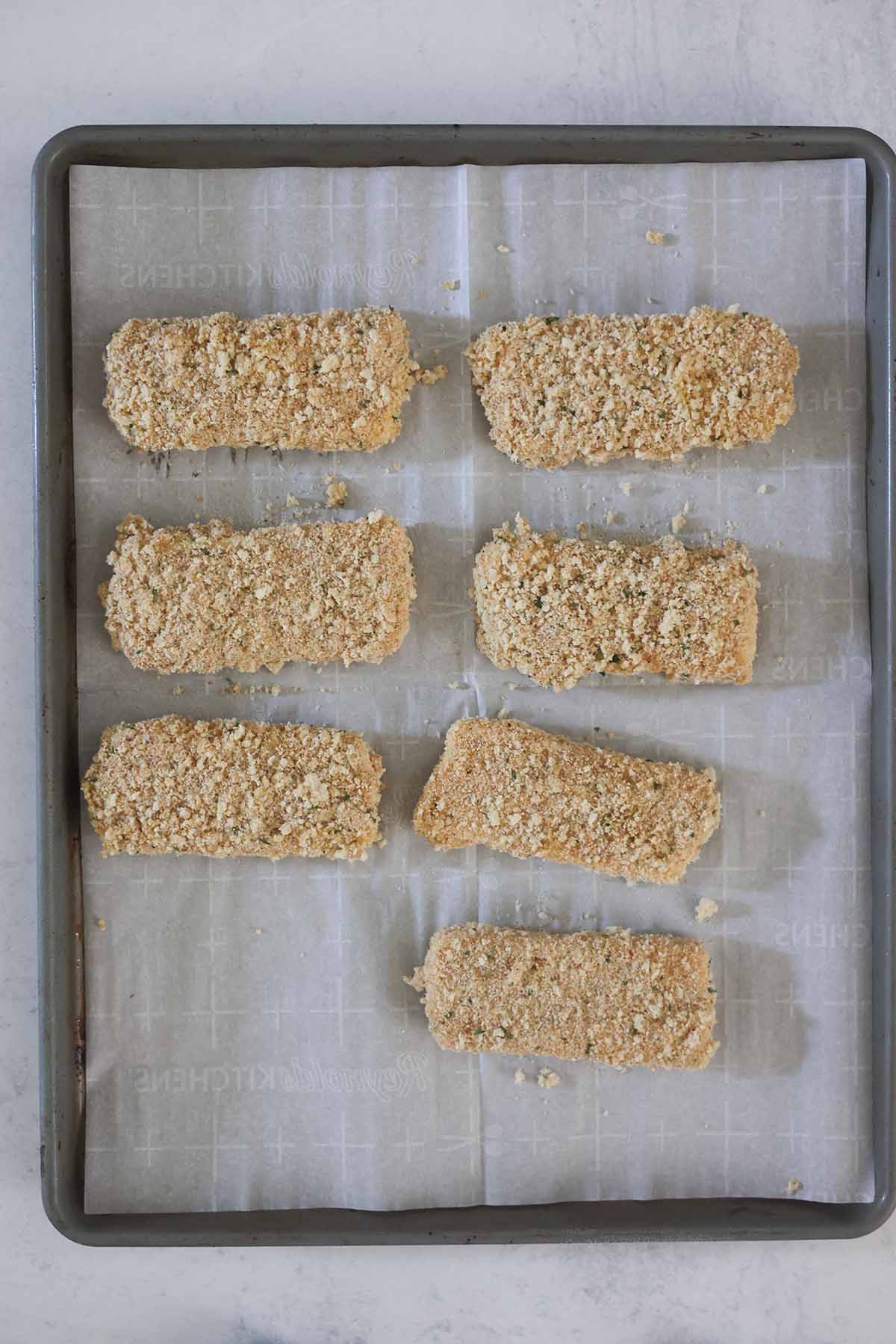 Seven bread crumb coated mozzarella sticks on a baking sheet.