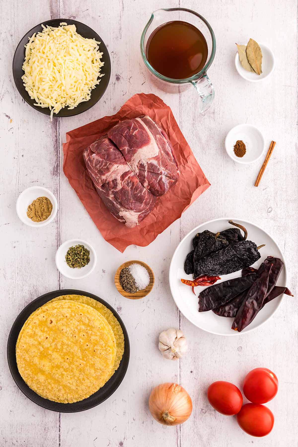Shredded cheese, beef broth, spices, bay leaves, raw beef, tortillas, tomatoes, onions, garlic, and cinnamon on a white surface.