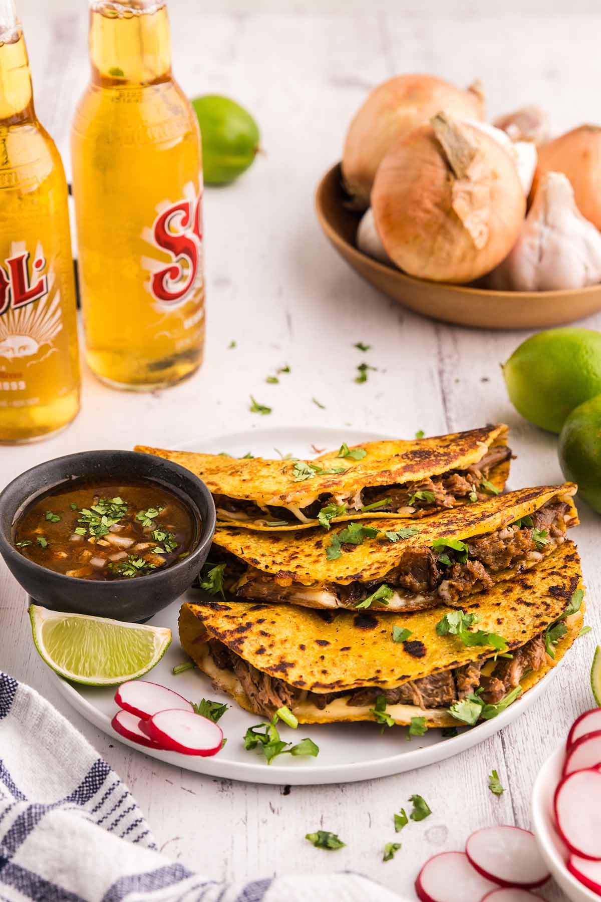 Three birria tacos with consome, limes, and sodas on the side.