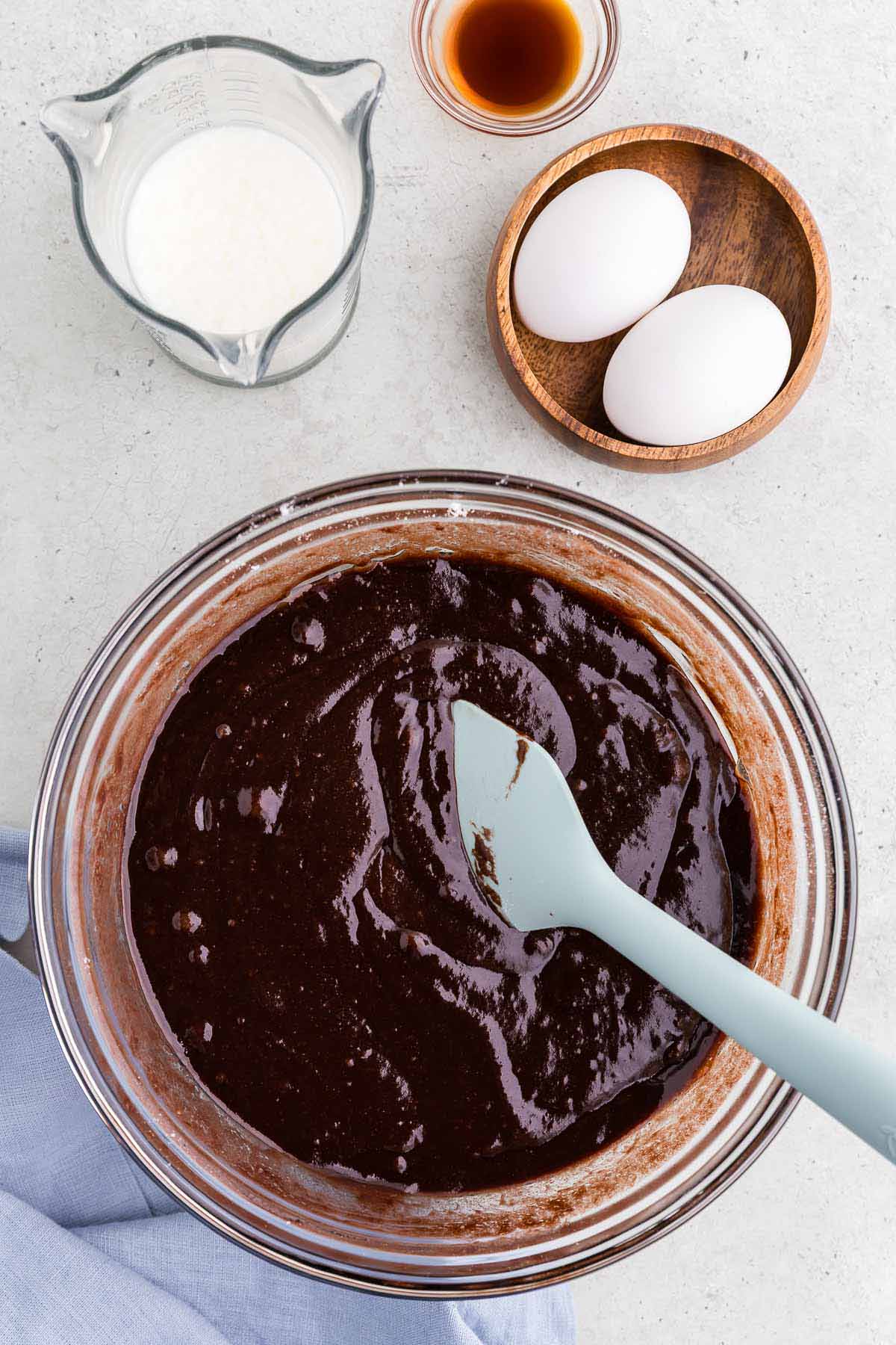 Chocolate cake batter in a bowl with eggs, milk, and vanilla extract on the side.