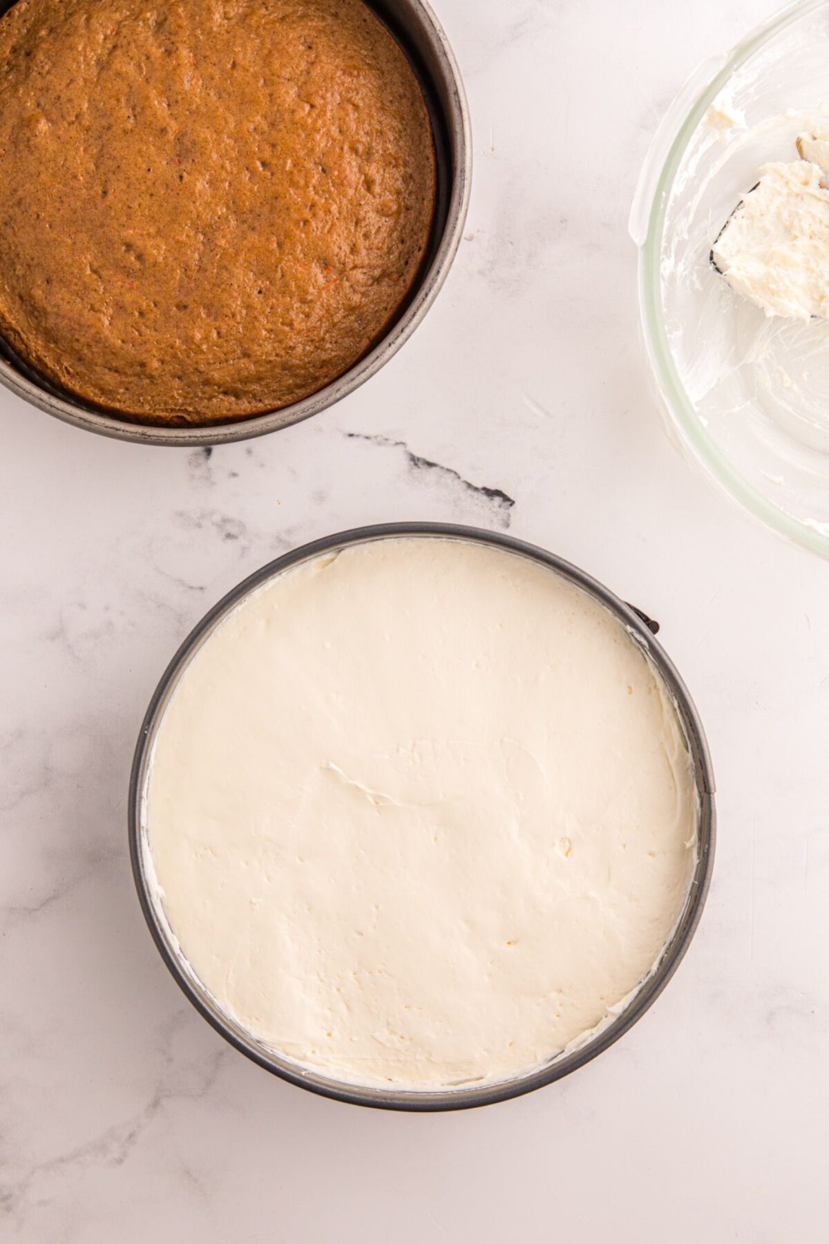 Baked cheesecake layer in a springform pan with carrot cake layer and cream cheese frosting on the side.