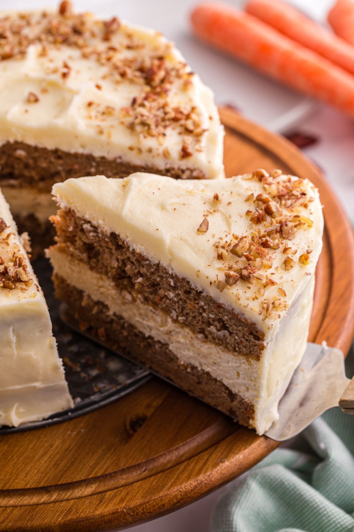 Layered carrot cake cheesecake with cream cheese frosting and chopped pecans on a wooden cake stand.