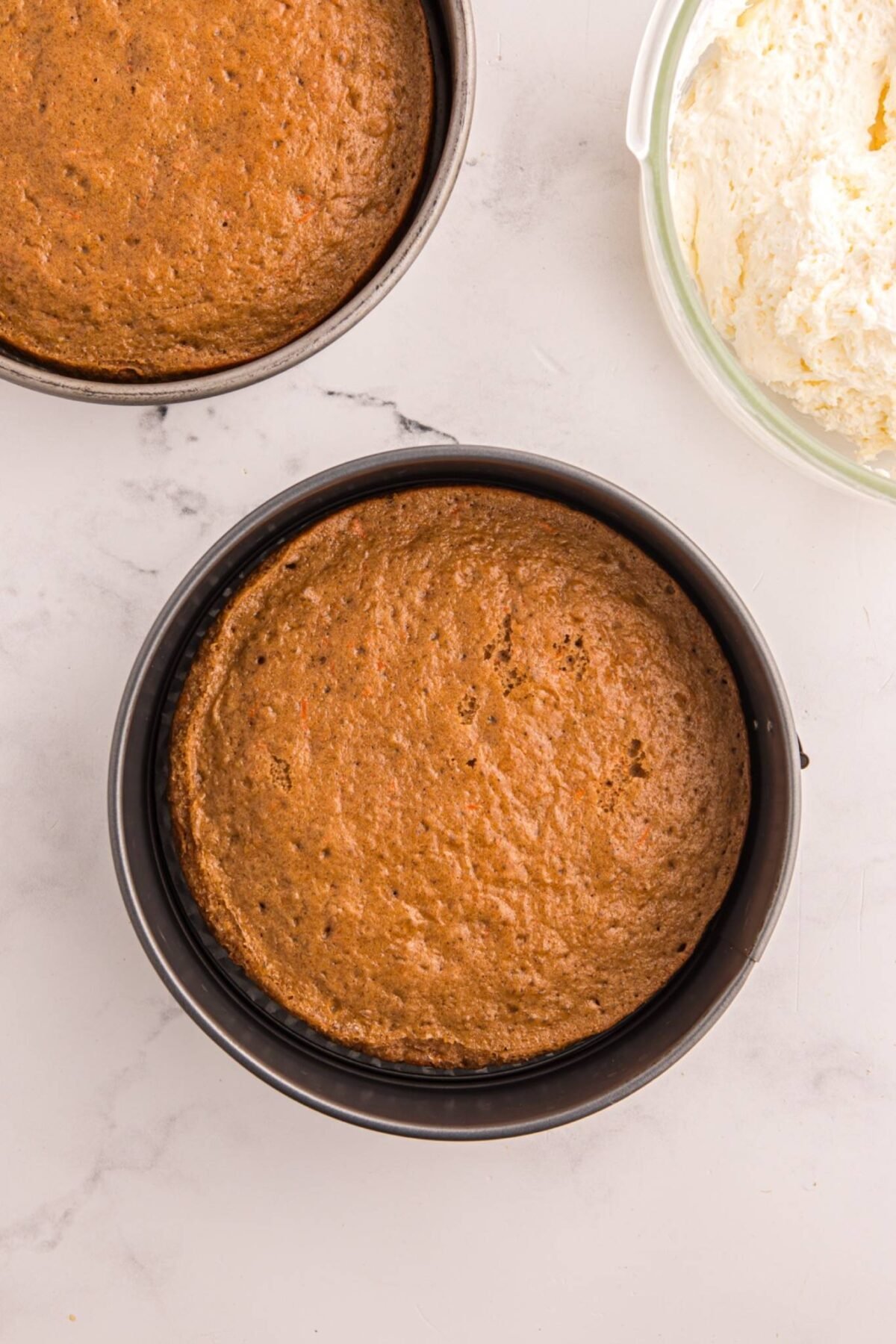 Two baked carrot cake layers in round cake pans with cream cheese frosting in a bowl.