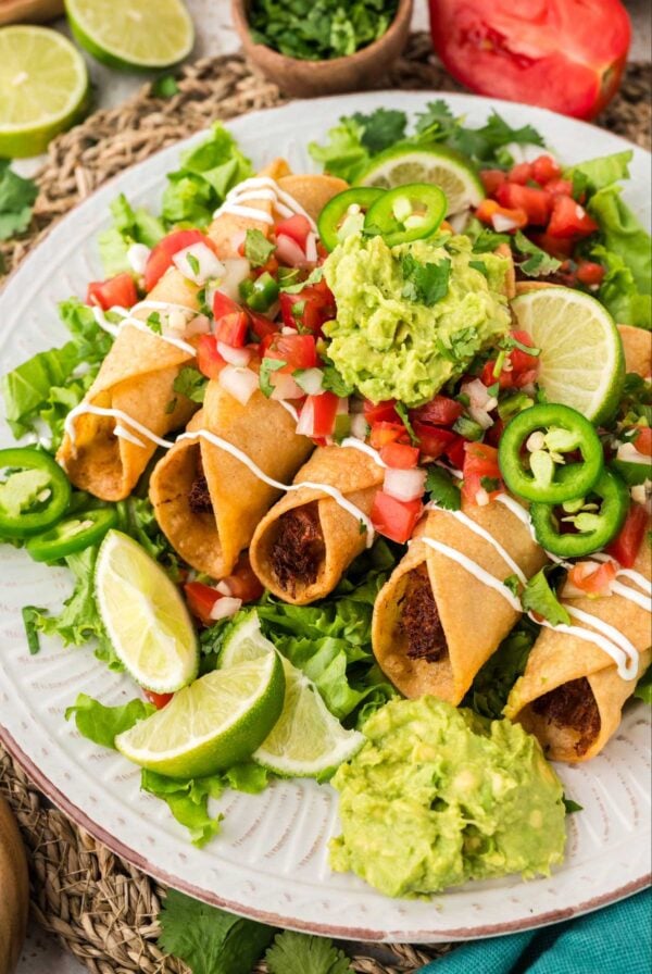 Chicken flautas on a plate with guacamole, pico de Gallo, jalapeños, and lime wedges,
