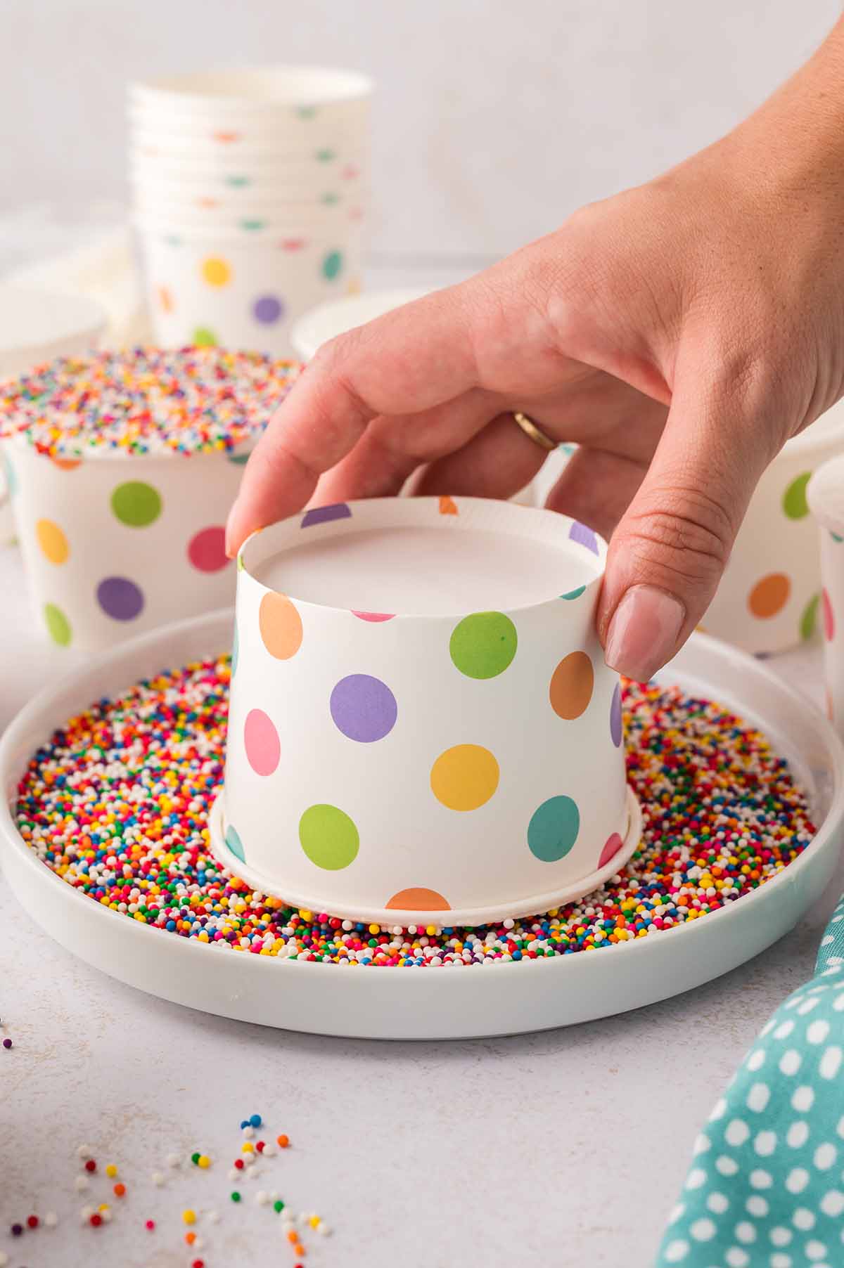Hand pressing a polka-dot cake cup into rainbow nonpareil sprinkles.