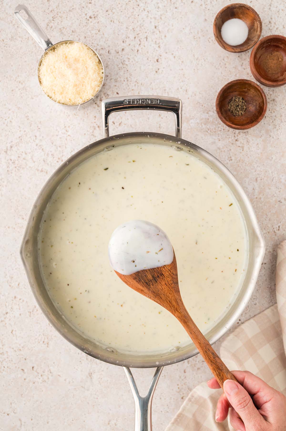 White sauce coating the back of a wooden spoon, with parmesan cheese and seasonings beside the skillet.