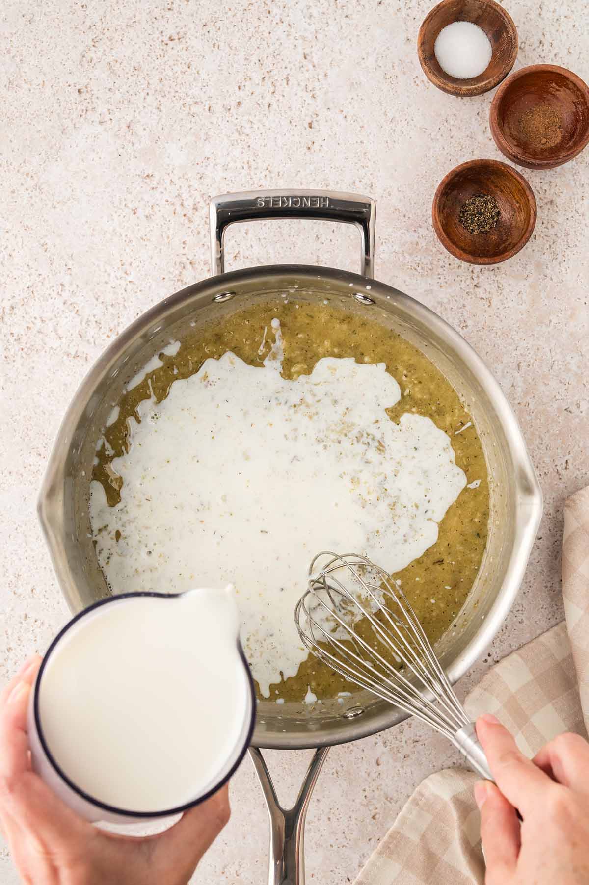 Hand pouring milk into a skillet with butter and flour while whisking to make a béchamel sauce.