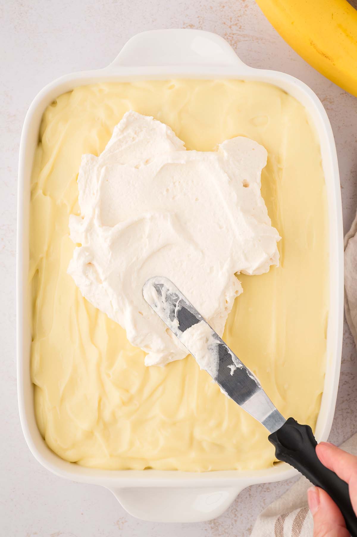 Whipped cream being evenly spread over the banana pudding.