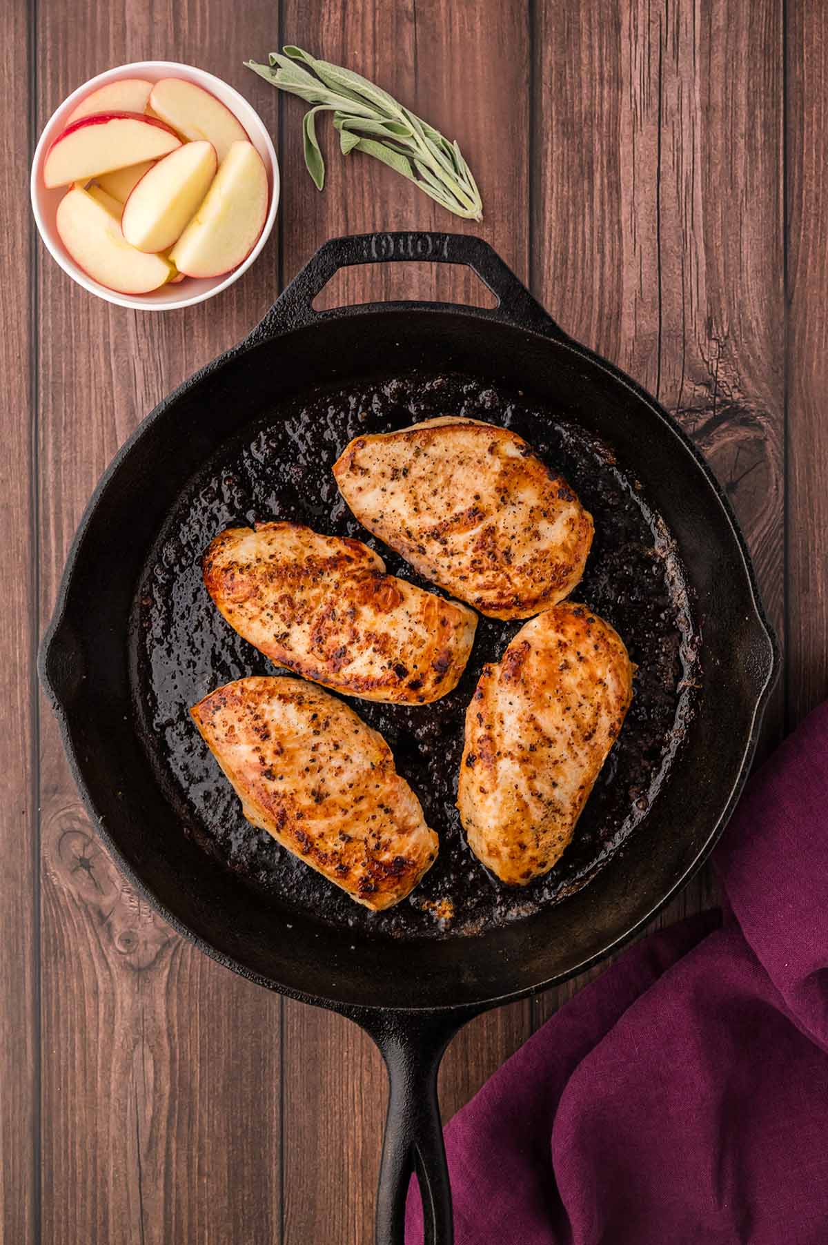 chicken cooking in a skillet with apples and sage on the side.