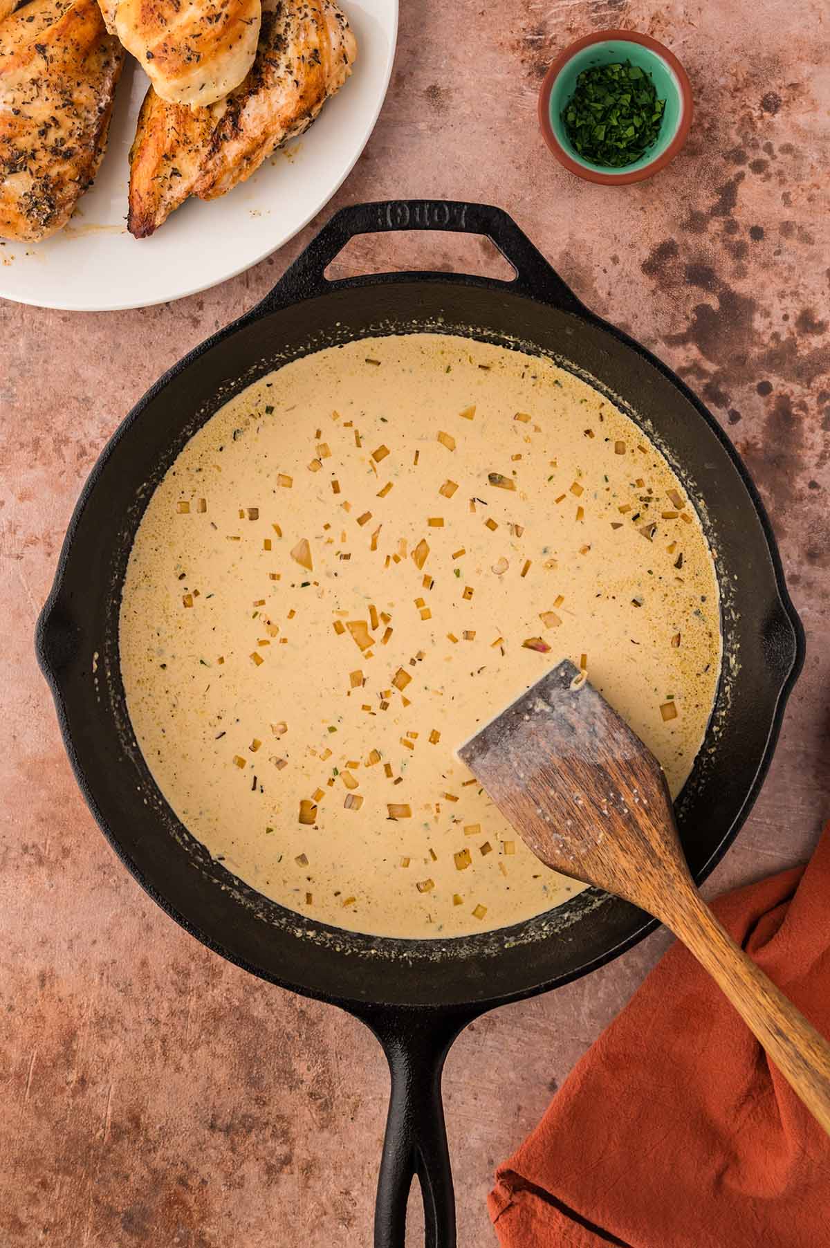 cream sauce in a skillet with a wooden spoon and chicken and parsley on the side.