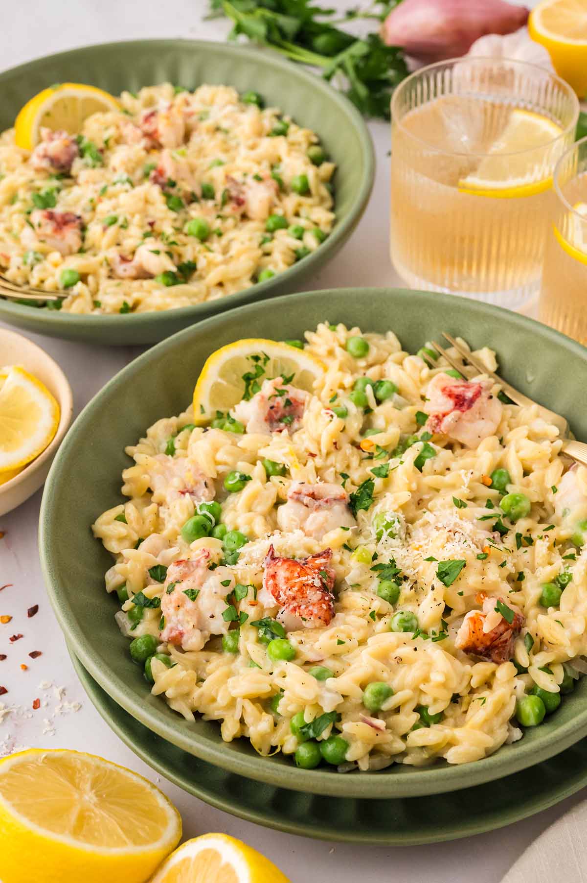 two green bowls filled with lobster orzo and lemon wedges and drinks on the side.