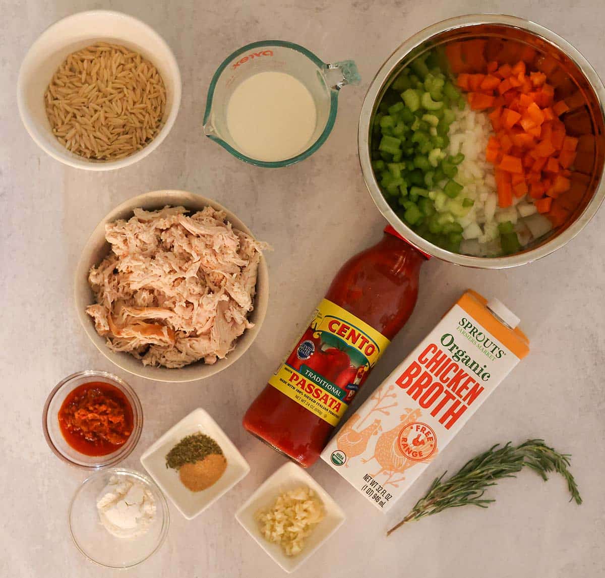 orzo, cream, veggies, shredded chicken, passata, broth, spices, rosemary, garlic flour, and chilies on a gray surface.