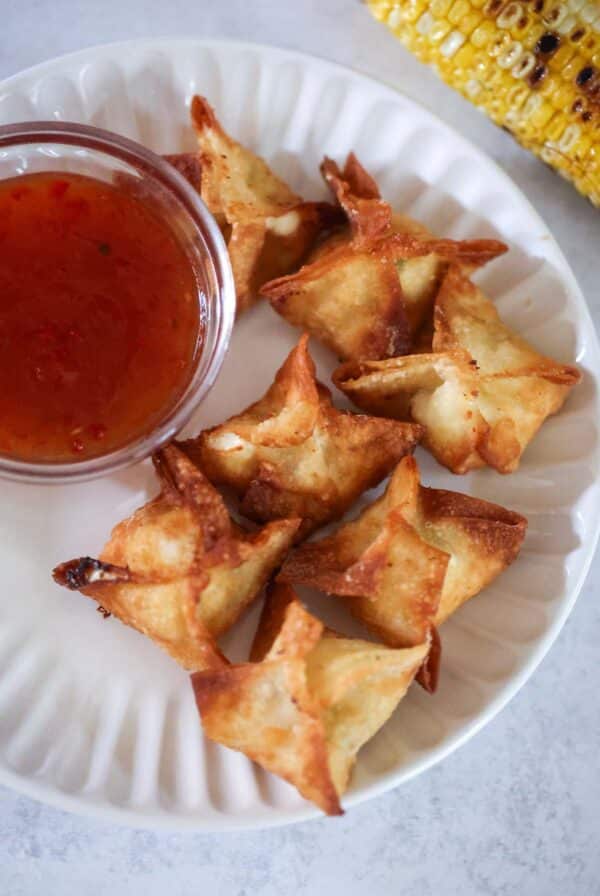 seven corn Rangoons on a white plate with sweet chili sauce on the side.