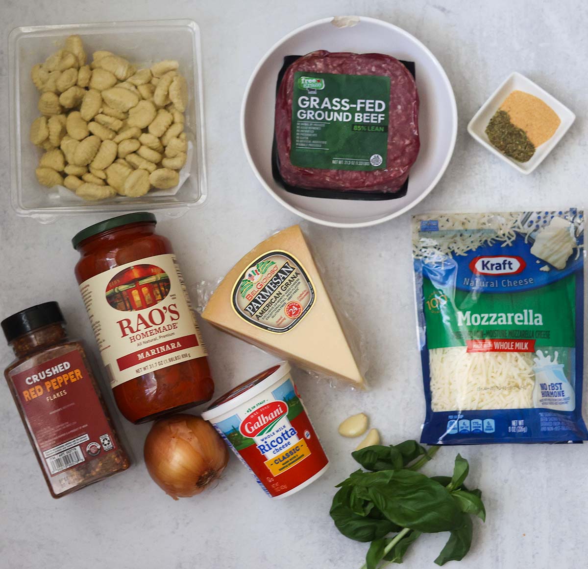 fresh gnocchi, ground beef, marinara sauce, cheese, spices, onions, garlic, and basil on a gray surface.