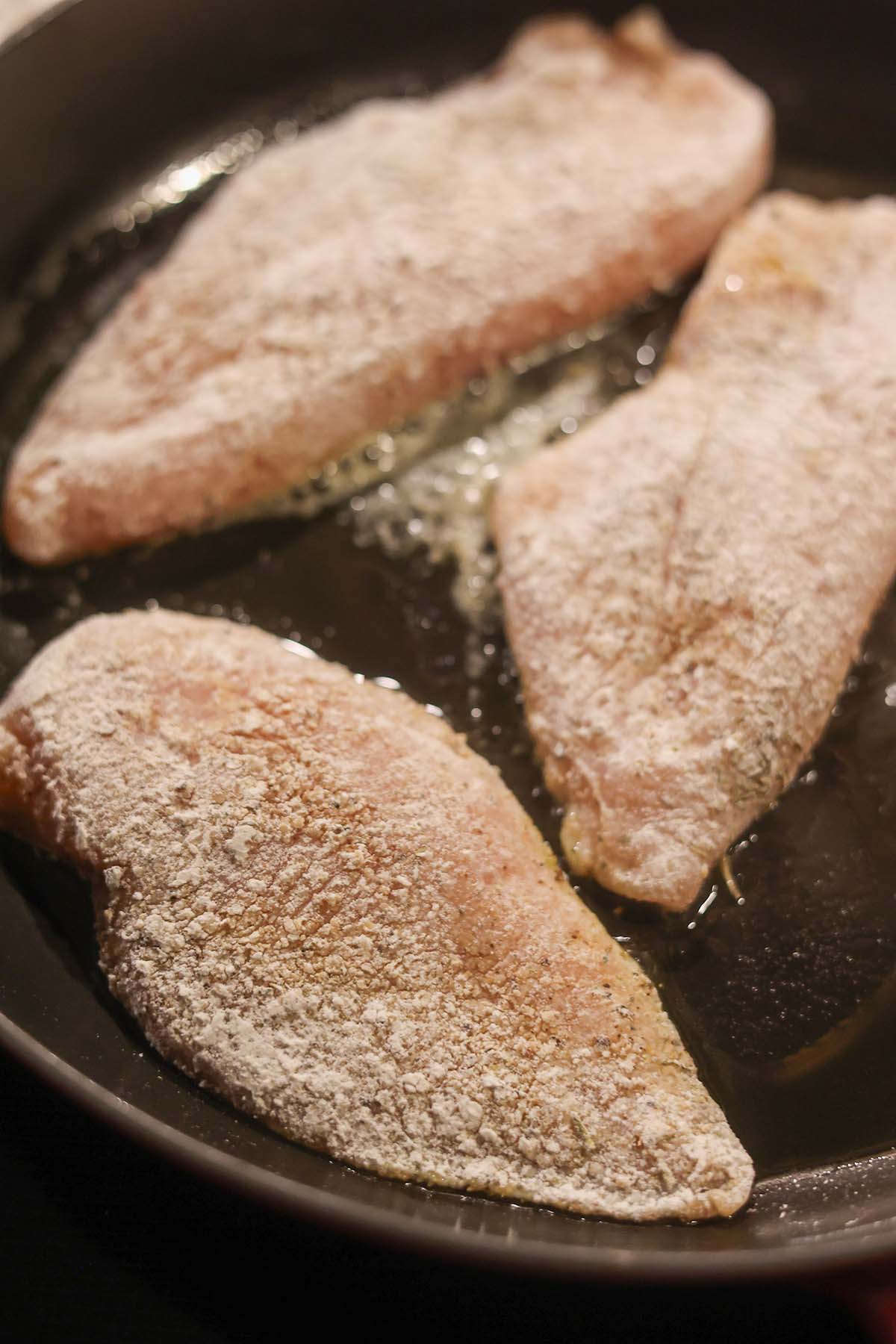 flour coated chicken breasts cooking in a skillet with oil.
